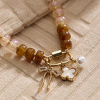 Textured Glass Bead Necklace with Charm Trio by Lilla Haven, featuring brown and cream glass beads accented with gold-toned spacers, and adorned with a trio of gold charms: a rhinestone-studded bow, a white flower, and a pearl—all elegantly displayed on textured beige fabric.
