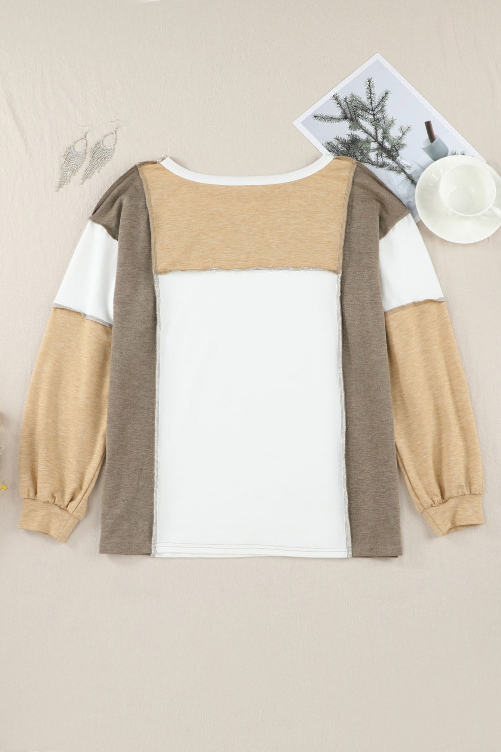 The Shewin Khaki Color Block Exposed Seam Long Sleeve Top, showcasing versatile khaki, beige, white, and brown tones, is laid flat on a beige surface. The background includes a pair of earrings, a magazine with a tree image, and a teacup with saucer. The unique patchwork design enhances its stylish appeal.