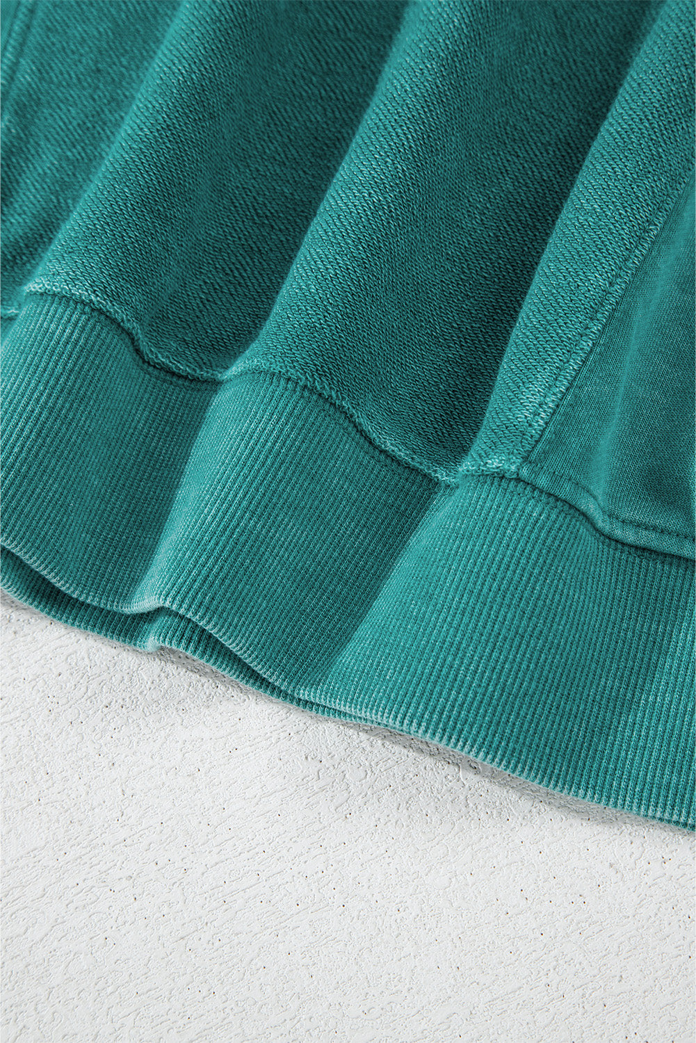 Close-up of the Shewin Notched Neck Exposed Seam Drop Shoulder Sweatshirt in green ribbed fabric, highlighting its textured hem on a flat surface.