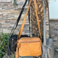 Three Roma Leathers, Inc. Cowhide Leather Crossbody bags with adjustable straps in black, tan, and gray hang from a palm tree outdoors, with a stone wall and greenery in the background.