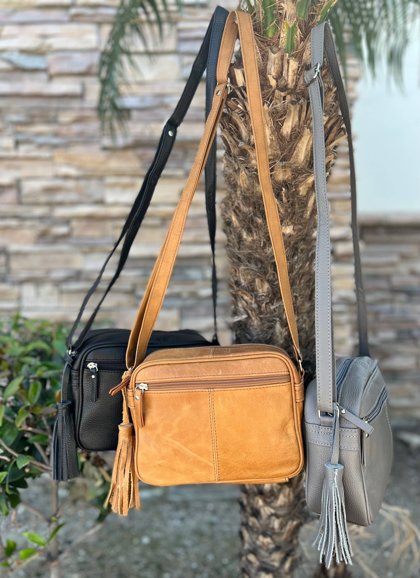 Three Roma Leathers, Inc. Cowhide Leather Crossbody bags with adjustable straps in black, tan, and gray hang from a palm tree outdoors, with a stone wall and greenery in the background.