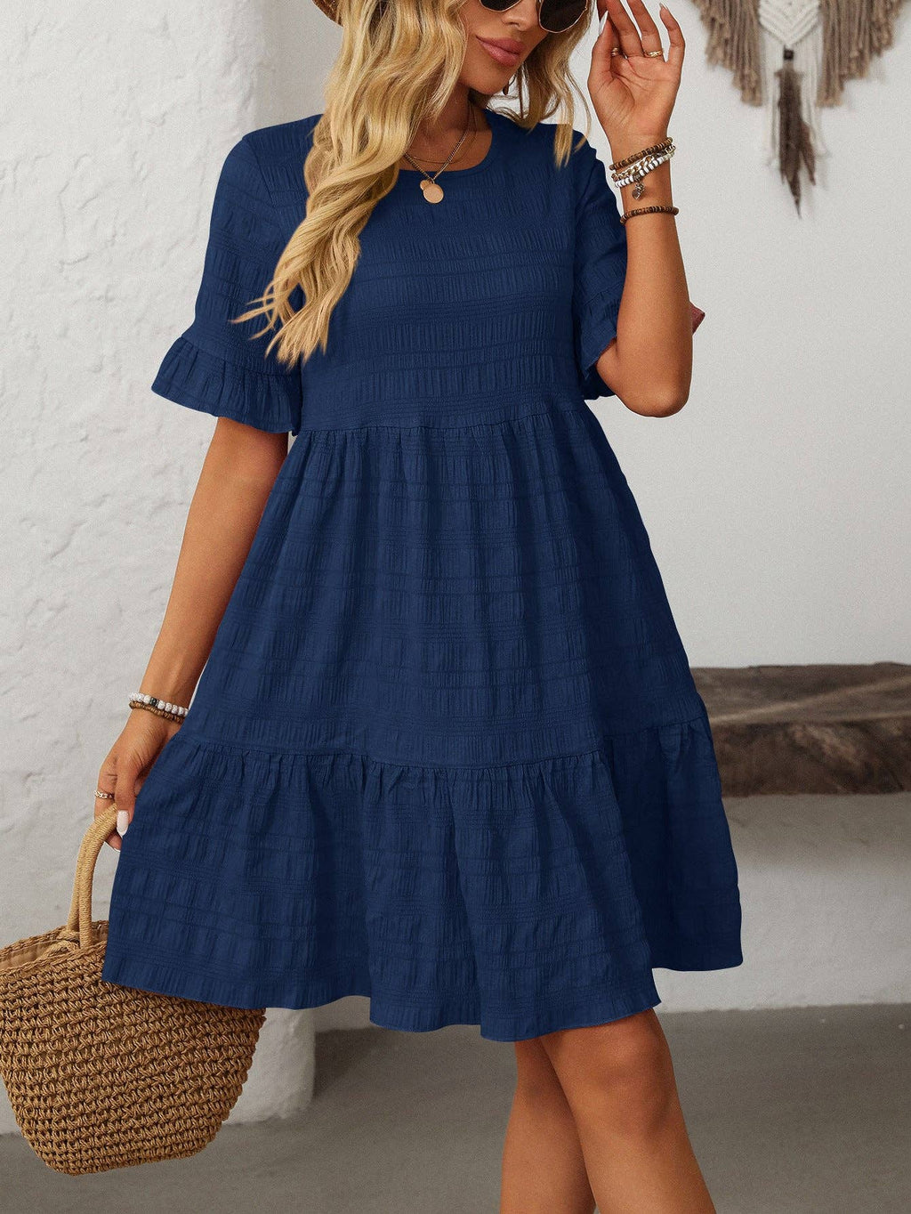 A woman stands indoors against a white wall, wearing the Suhkasana Loose Casual Short-Sleeve Dress in elegant navy blue and holding a woven straw handbag.