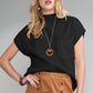 Against a neutral backdrop, the woman showcases a black top paired with a brown skirt and a necklace featuring a circular pendant. Perfect for transitional weather, her ensemble is complemented by Shewin's versatile Patch Pocket Ribbed Knit Short Sleeve Sweater.