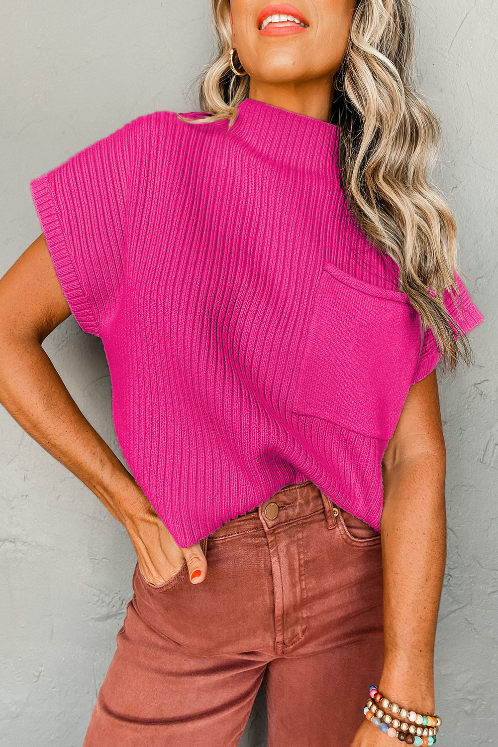 The Shewin Patch Pocket Ribbed Knit Short Sleeve Sweater in vibrant pink, featuring a mock neck and pocket, is an ideal transitional piece paired with high-waisted brown pants against a sleek gray background.