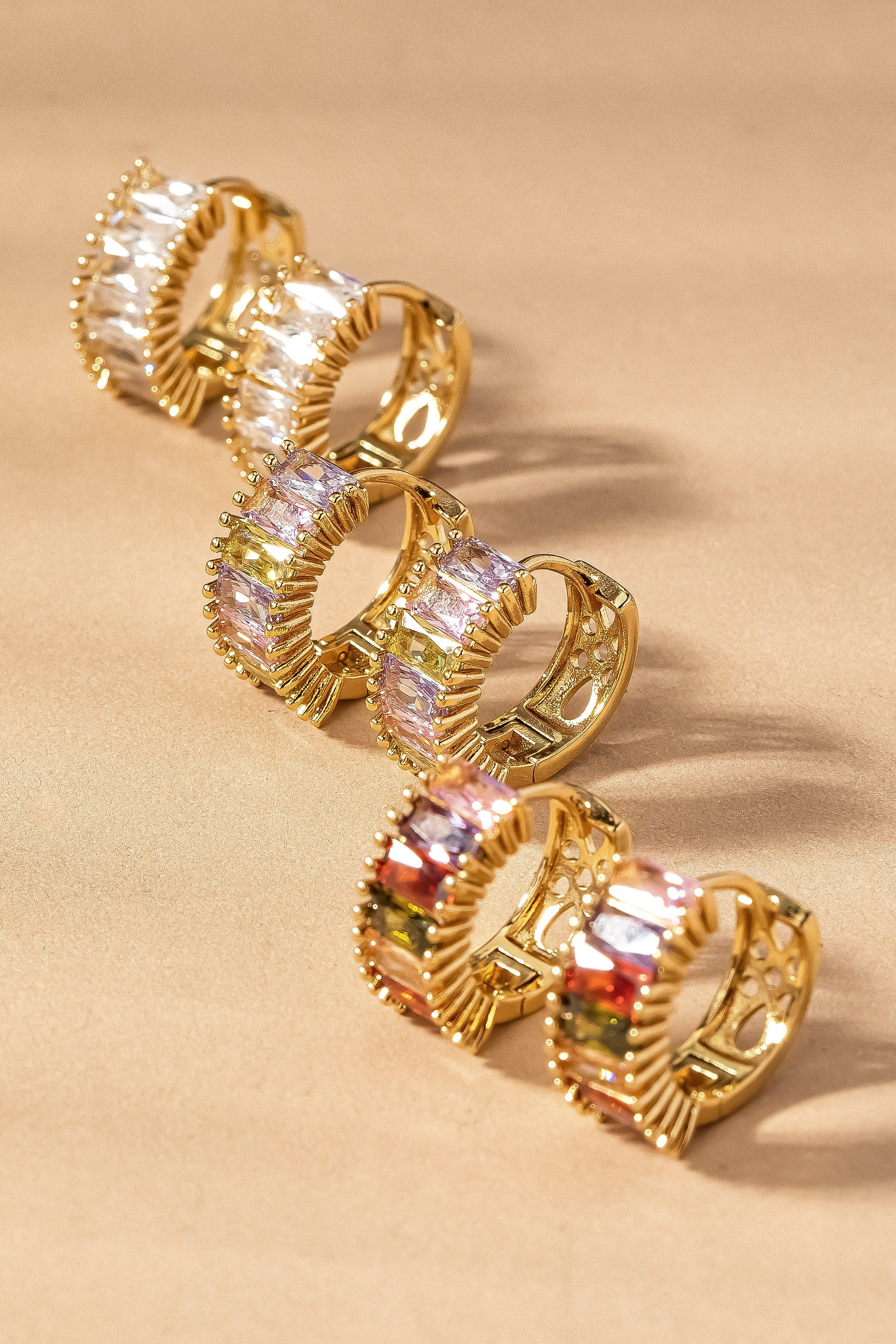 Several elegant gold rings with FASHION GO Huggie with Rectangular CZ earrings arranged in a line on a textured beige background, reflecting light. Main image