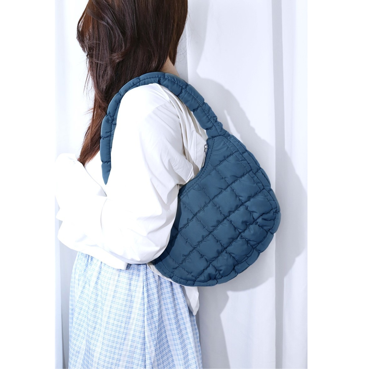 A person with long hair, wearing a white blouse and a blue plaid skirt, carries the Zenana Puffer Quilted Mini Bag in assorted colors with a comfortable shoulder strap over their shoulder.
