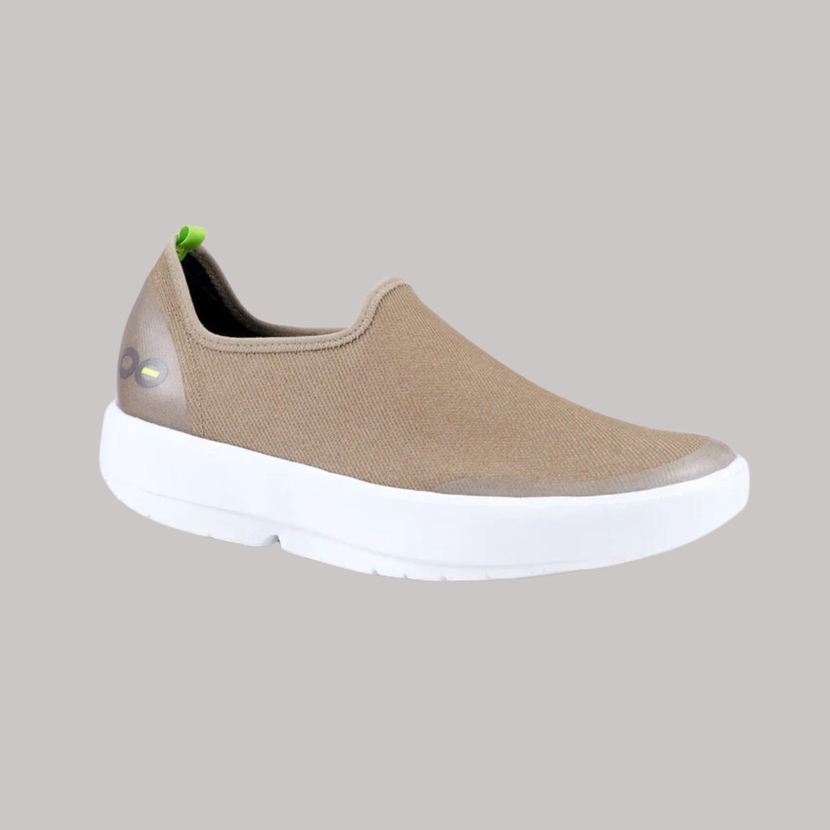 The OOFOS OOMG EASY LOW TAUPE, from OOFOS LLC, offers ultimate comfort with its 4-way stretch design and is showcased with a stylish white sole and a small green tag at the heel against a simple gray background.