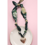 The FASHION GO Handmade Scarf Necklace, shown in a green patterned fabric with assorted charms including a gold heart-shaped locket, is displayed on a white plate against a pink background.