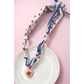 A FASHION GO Handmade Scarf Necklace in assorted colors and patterns is styled on a gold-rimmed plate, showcasing its trendy heart pendant with red center. The setup features a pink background.