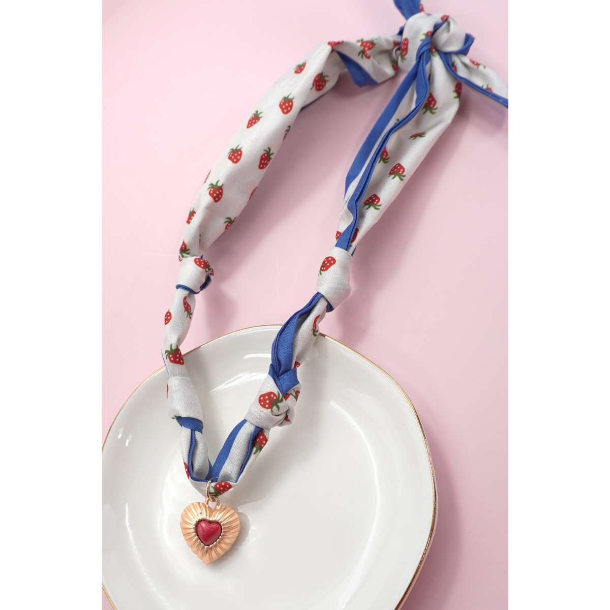 A FASHION GO Handmade Scarf Necklace in assorted colors and patterns is styled on a gold-rimmed plate, showcasing its trendy heart pendant with red center. The setup features a pink background.