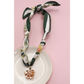 The FASHION GO Handmade Scarf Necklace in assorted colors and patterns, adorned with charms and a gold pendant with pearl accents, is showcased on a white dish against a pink background.