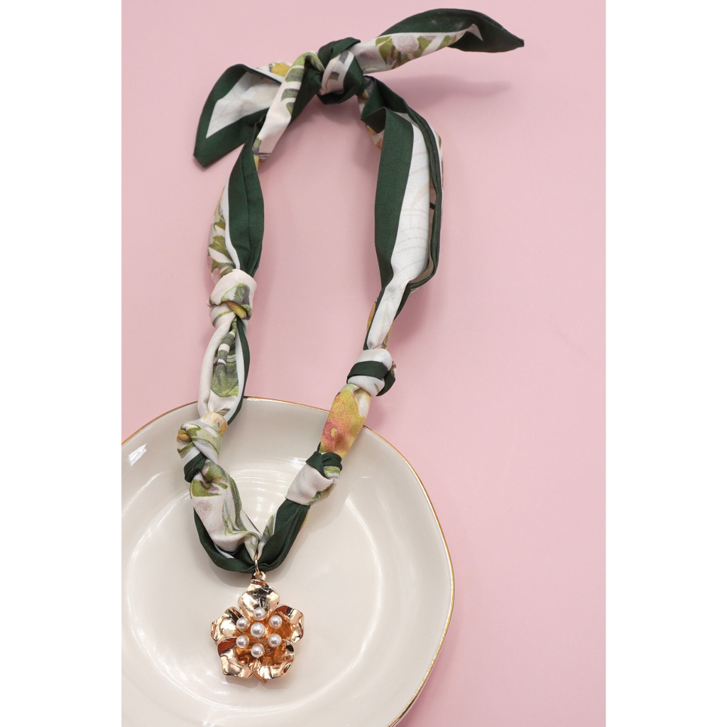 The FASHION GO Handmade Scarf Necklace in assorted colors and patterns, adorned with charms and a gold pendant with pearl accents, is showcased on a white dish against a pink background.