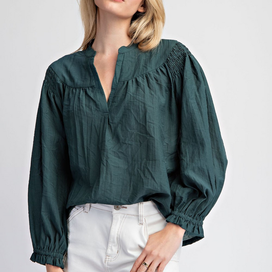 A person wearing the Crinkled Notched Neck Long Sleeve Blouse in Hunter Green from FASHION GO and white pants, with blonde hair partially covering their face.