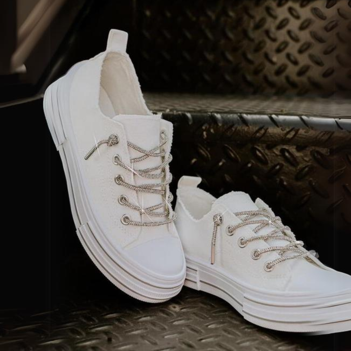 The Aman Tennis Shoe in White by VERY G, featuring a canvas upper and rhinestone laces, is displayed on a black, textured metal staircase.