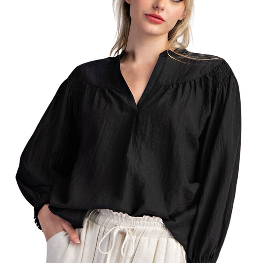 A woman is wearing the Crinkle Notched Neck Long Sleeve Top in Black by FASHION GO paired with light-colored drawstring pants made from lightweight material. She has one hand in her pocket.