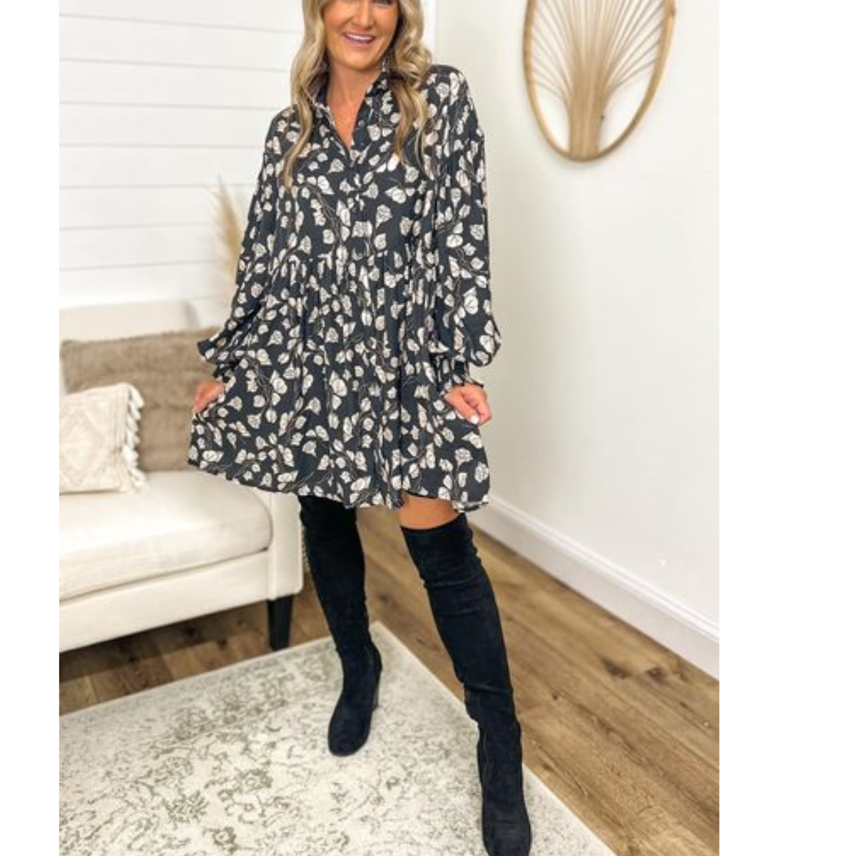 A woman in an Entro Floral Dress in Dark Charcoal and Ivory with black knee-high boots stands inside a room with light-colored walls, a white armchair, a light-colored rug, and a wall decoration behind her.