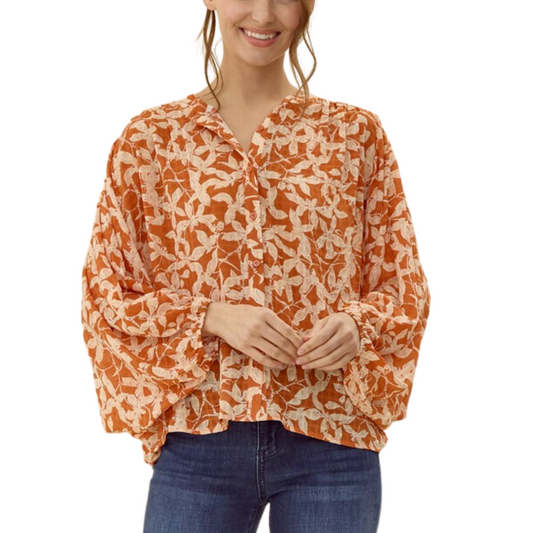 A woman wearing an Orange Floral Print Blouse with long dolman sleeves from FASHION GO and blue jeans stands with her hands lightly clasped.