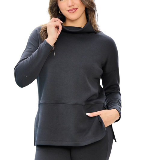 A woman is wearing the Vienna Top in Black by Dear Scarlett, with one hand touching her collar and the other in her pocket. The background is white, highlighting the elegant design of her cowl neck.