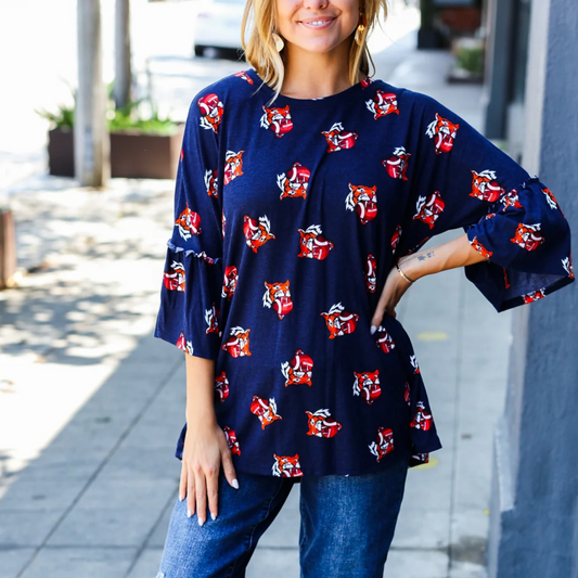 Person wearing a Tigers Gameday Top by Bloom Wholesale, featuring blue polyester with red and white fox prints, standing on a sidewalk with a hand resting on their hip.