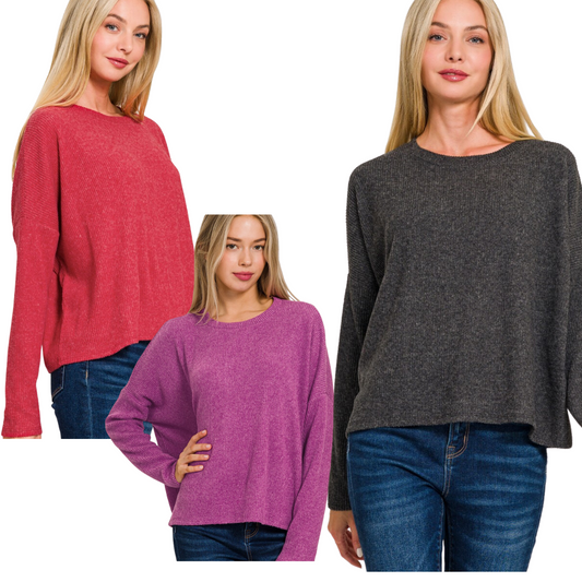 Three women are modeling Zenana Ribbed Dolman Long Sleeve Lightweight Sweaters. One wears a red sweater, another a pink sweater, and the third a gray one. They pair the sweaters with blue jeans and have straight, long, blonde hair.