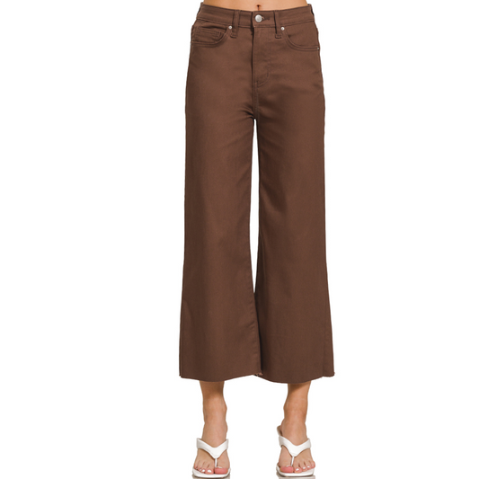 A person wearing Zenana's High Rise Crop Colored Pants in Brown paired with white sandals is photographed from the waist down. The ensemble blends the stylish appearance of high-rise crop pants with the casual ease of summer footwear.