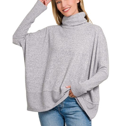 A person wears an oversized, light gray Brushed Mélange Hacci Turtleneck Sweater from Zenana, paired with blue jeans. They are smiling and holding the edge of the super soft Heather Grey sweater with one hand.