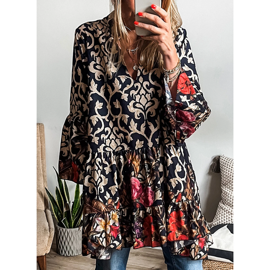 A person wearing a Shewin Black Abstract Print V-Neck Tiered Mini Dress featuring red and orange floral designs takes a mirror selfie. Flounce sleeves add a whimsical touch, while a plant and a woven pillow create an inviting background.