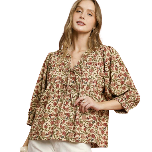 A woman wears the Umgee Vintage Floral Pintuck Blouse featuring a loose fit, V-neck, and lace-up detail, paired stylishly with white pants.