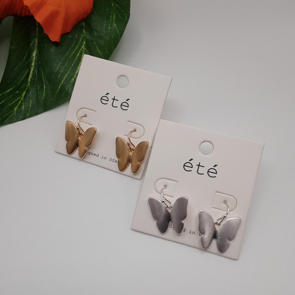 Two pairs of "Drop Butterfly Earring Matte Metal" by SPECIAL EFFECTS are displayed on white cards labeled “été.” One pair is gold-toned, and the other is silver-toned. A green leaf and an orange flower can be partially seen in the background.