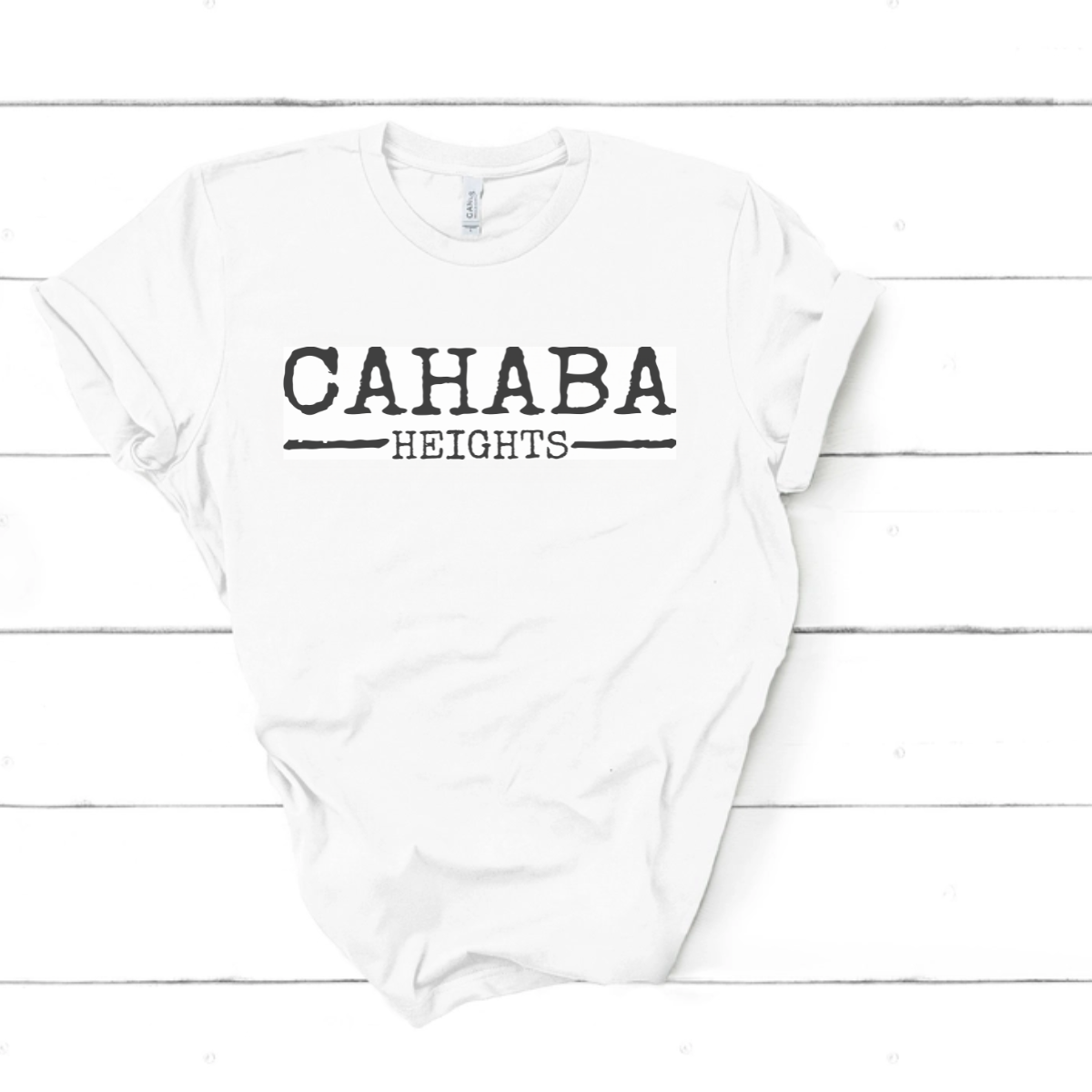 The Faire Distressed Front Cahaba Heights T-Shirt features "CAHABA HEIGHTS" in bold black text, shown laid flat on a white wooden surface.