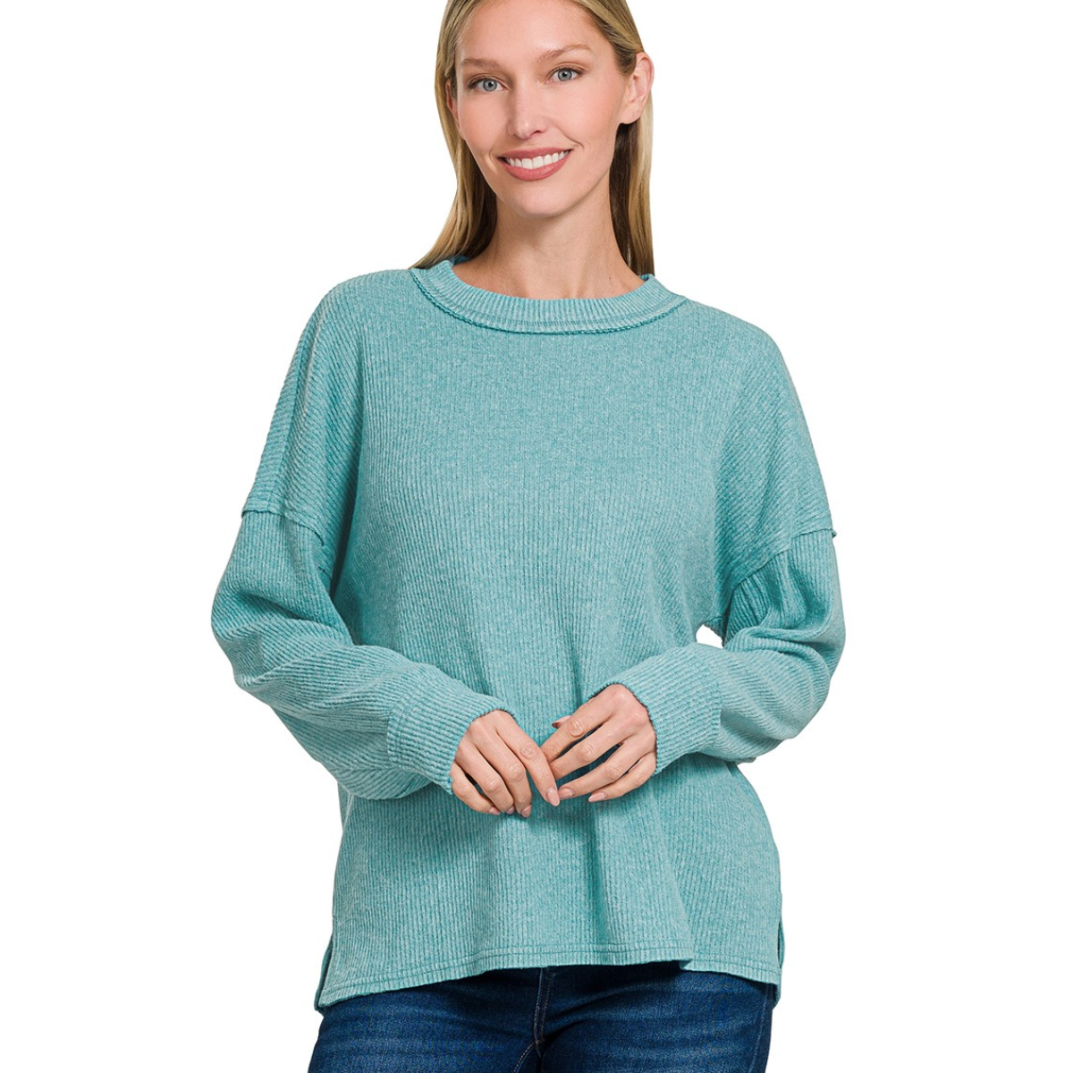 A woman stands facing the camera, wearing the Zenana Brushed Ribbed Hacci Drop Shoulder Sweater in light teal with blue jeans, and smiles gently.