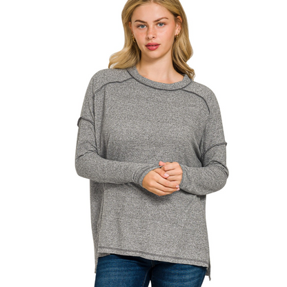 A woman with long blonde hair models the Zenana Soft Melange Hacci Exposed Seam Hi-low Tee, a loose-fitting gray long-sleeve sweater, paired with blue jeans against a plain white background.