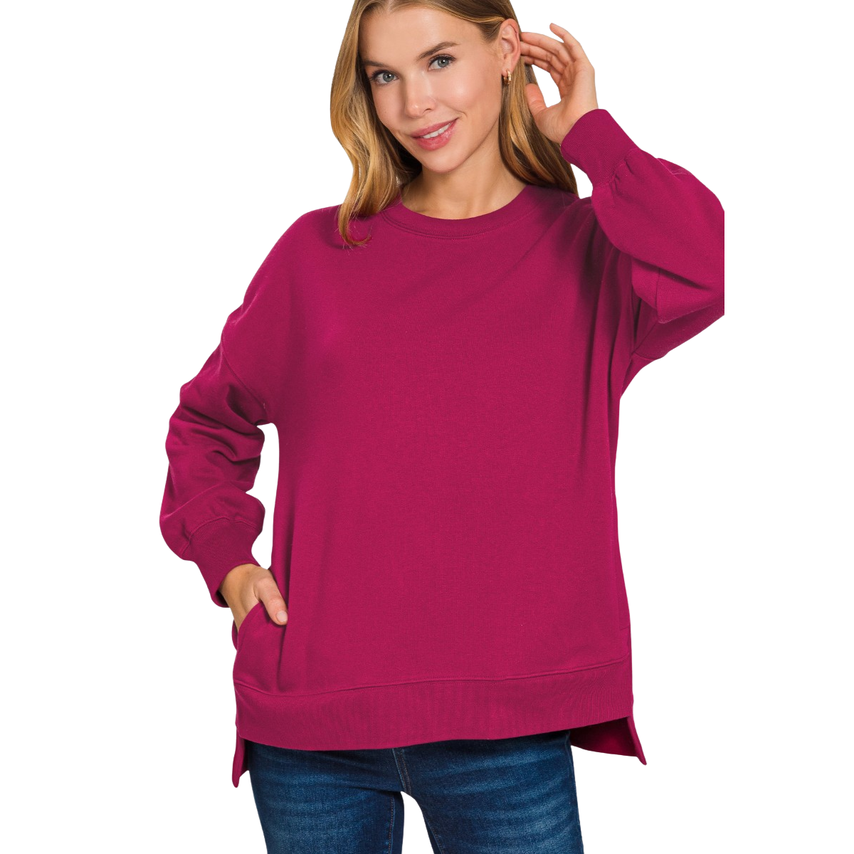 A woman models the Zenana Fleece Hi-Low Hem sweatshirt in bright pink with blue jeans, one hand in her pocket and the other touching her hair.