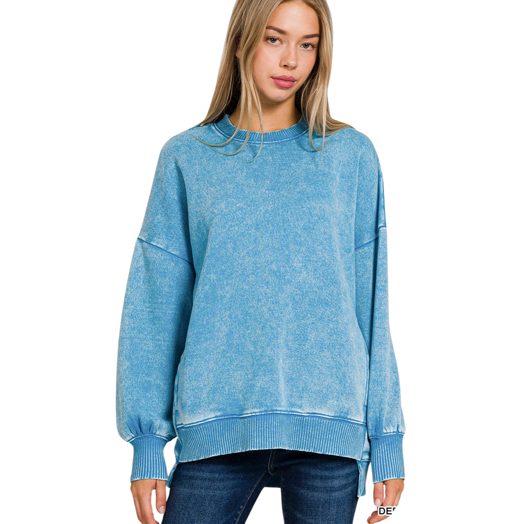 A young woman with long blonde hair models the FASHION GO Acid Washed Fleece Hi-Low Pullover in light blue, paired with dark jeans, standing against a white background.