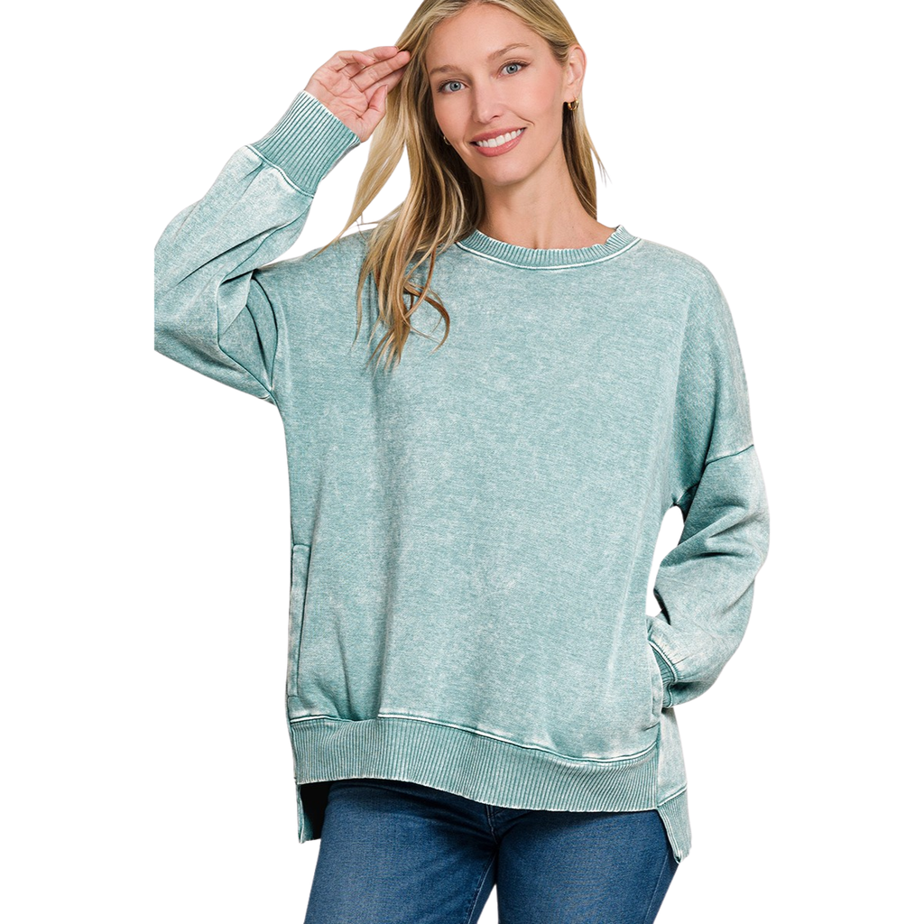 A woman smiles at the camera wearing the FASHION GO Acid Washed Fleece Hi-Low Pullover in light teal with blue jeans, standing with one hand in her pocket and the other touching her hair.