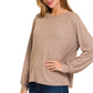 A woman wearing a Zenana Soft Melange Hacci Round Neck Long Sleeve Tee in beige and blue jeans stands with one hand on her hip, facing slightly to the side.