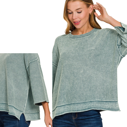 A woman wears the Zenana Washed 3/4 Sleeve Boxy Cotton Sweater, a loose-fitting, light teal 100% cotton top with exposed seams and layered hem, styled with blue jeans. Close-up highlights sleeve detail on her left arm.