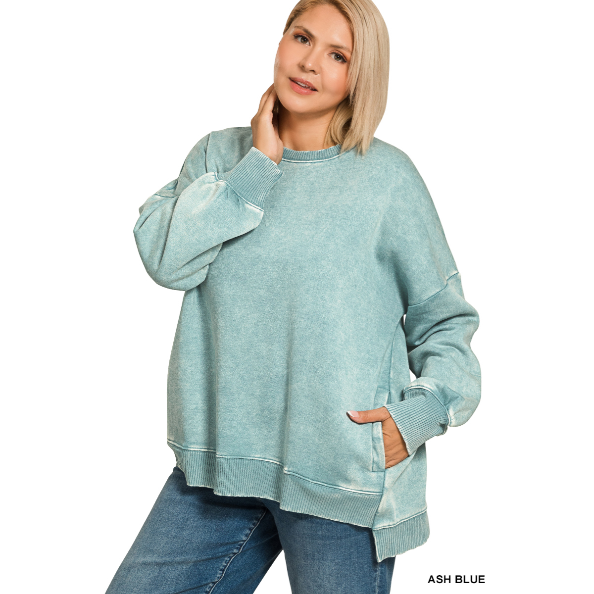 A woman models the FASHION GO Plus Acid Washed Fleece Hi-Low Hem Pullover in ash blue with blue jeans, posing against a white background with one hand on her neck and the other in her pocket. Main image