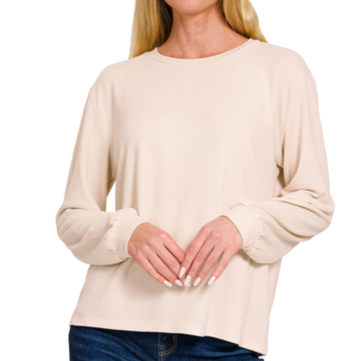 A woman wearing the Zenana Soft Melange Hacci Round Neck Long Sleeve Tee in beige and blue jeans stands with her hands folded in front.