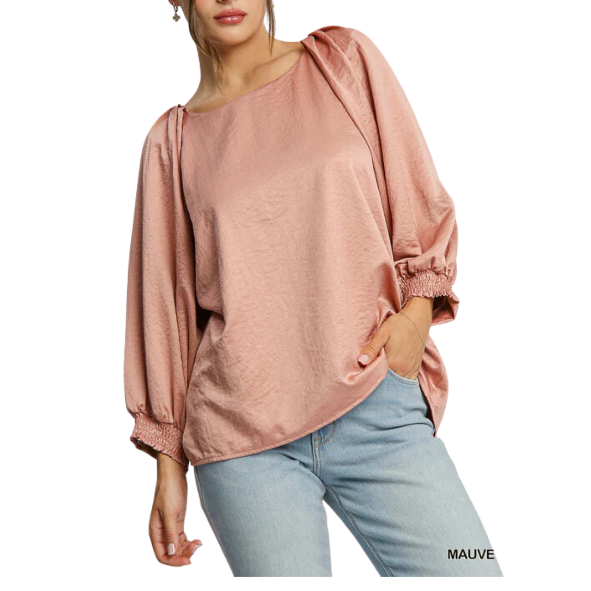 A person is wearing the Umgee Round Neck High Low Top in a loose, long-sleeved style with smocked cuffed sleeves paired with light blue jeans, featuring a back button keyhole for added elegance, posed gracefully with one hand in the pocket.