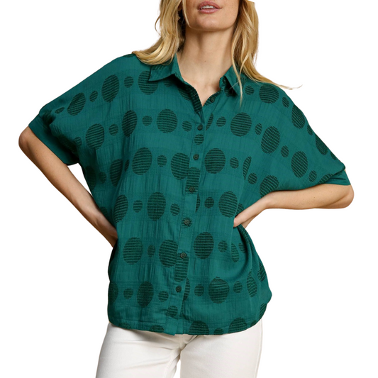 A woman stands with her hands on her hips, wearing white pants and a green Textured Polka Oversized Top in Peacock by Umgee.