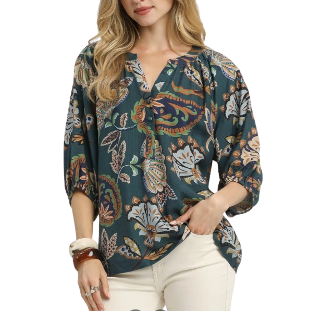 A woman wears the Umgee Boho Paisley Puff Sleeve Blouse paired with cream pants and bracelets, featuring a teal paisley print, split neckline, and three-quarter puff sleeves, standing with one hand in her pocket.