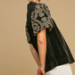 A person is wearing the Velvet Embroidered Sleeve Top in Black by Umgee, featuring intricate embroidered floral patterns on the bubble sleeves. They are shown from the side against a plain beige background.