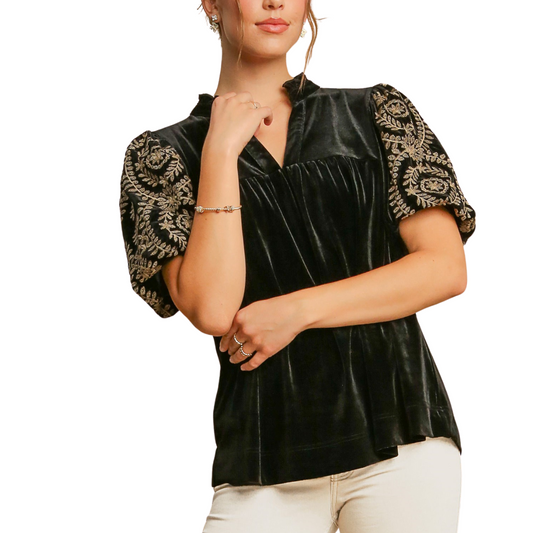 A person wearing the Velvet Embroidered Sleeve Top in Black by Umgee, featuring ornate designs on the sleeves and a ruffle split neck, poses with one hand near their chin against a white background.