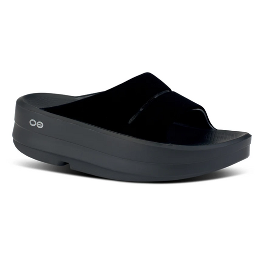 The OOMEGA OOahh Black Posh sandal by OOFOS LLC features a thick sole with superior shock-absorbing comfort, a single wide strap over the foot, and a small "OO" logo on the side.