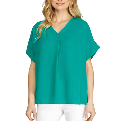 A person stands against a white background wearing a teal V-Neck Short Sleeve Air Flow Blouse by She & Sky, paired with white pants.