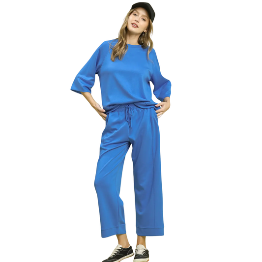 A woman wears the Umgee 2 Piece Activewear Set—blue, with a loose drop shoulder top and elastic waist pants—paired with black sneakers and a black cap, standing hands on hips.