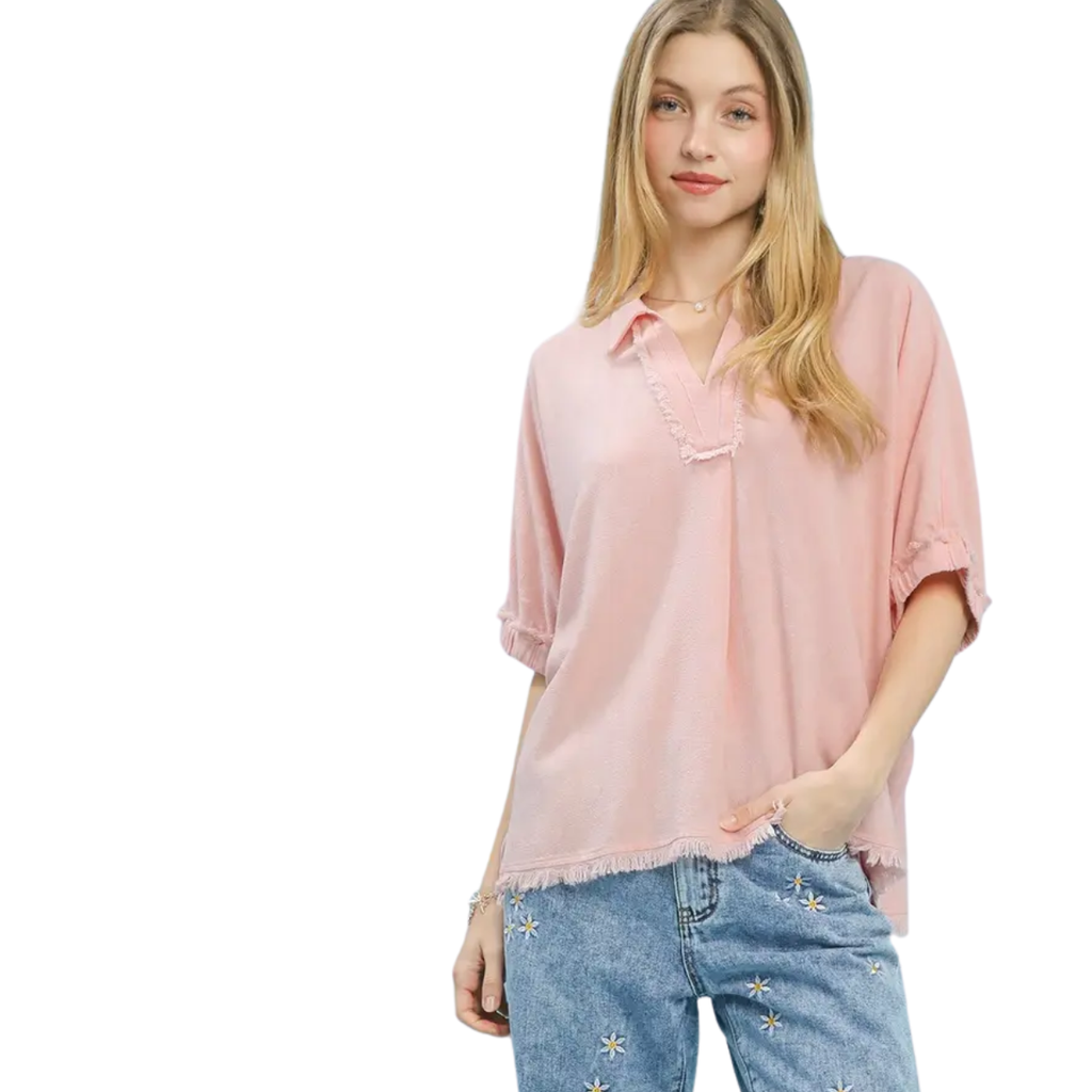 A woman wears the Umgee Linen Frayed Edge Split Neck Short Sleeve Top in light pink, styled with blue embroidered jeans. She stands with one hand in her pocket against a white background.