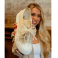 A woman holds the VERY G Aman Sneaker with Pearl and Sparkle Laces—a beige shoe featuring white laces, a sheer bow, and pearl accents—against a blurred brick wall background.