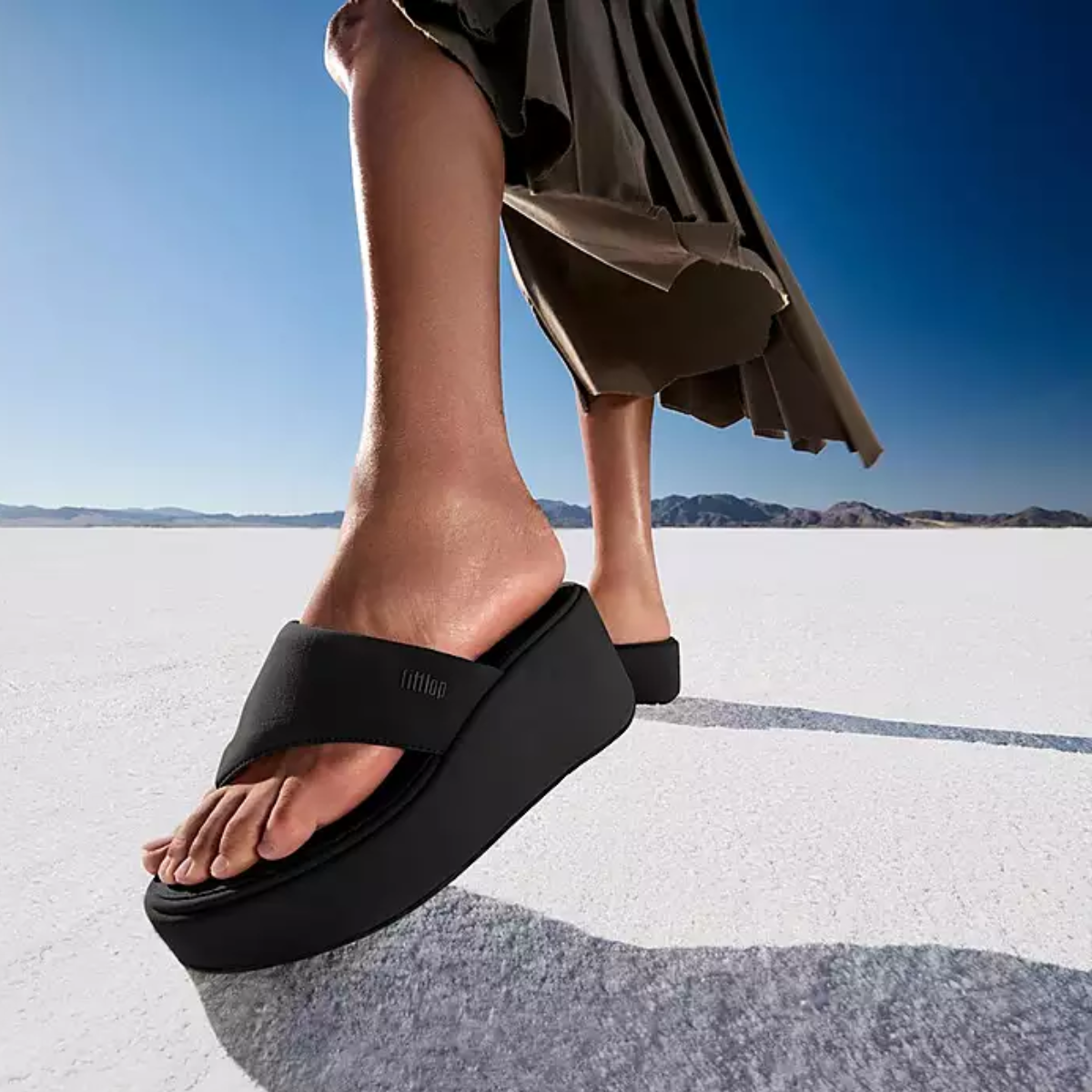 A person in a dark skirt walks outdoors on a flat, white surface with mountains behind, wearing fit flop Platfforms Wedge Flip-Flops - Black under a clear blue sky.